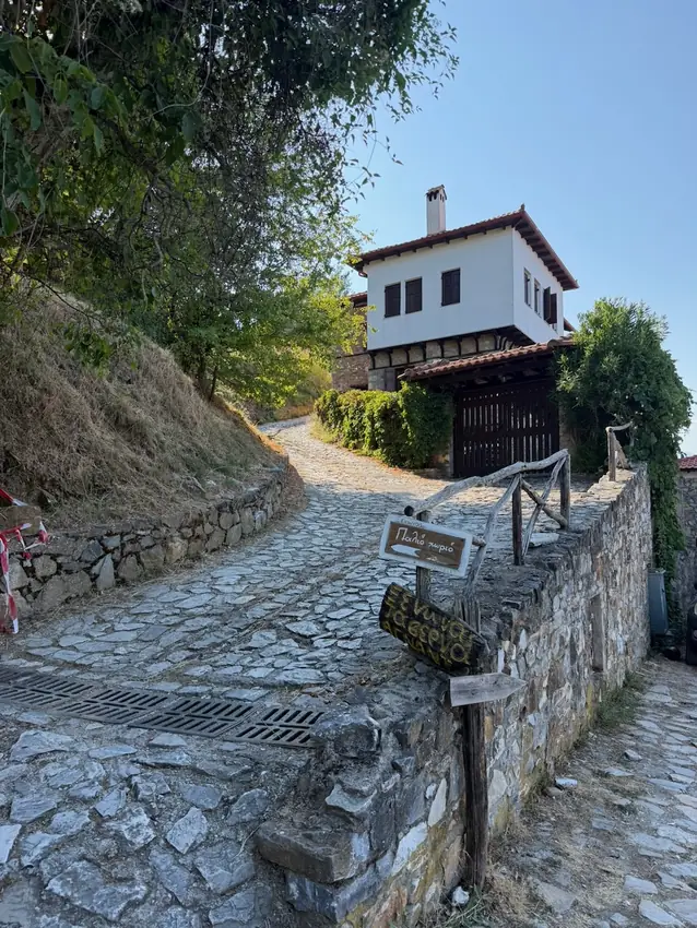 Cobbled road up to the old centre of Palaios Panteleimonas with a white stone house
                    and a signpost