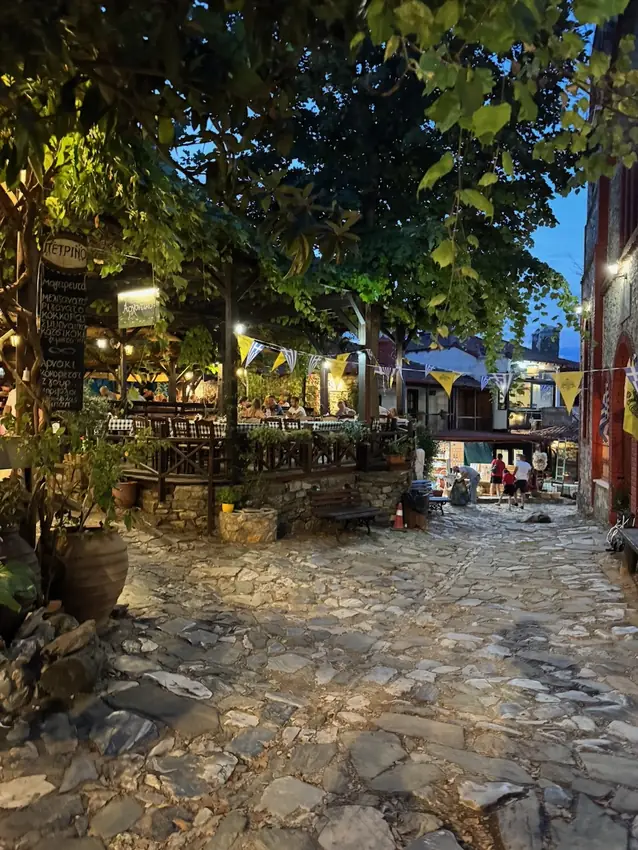 The village square of Palaios Panteleimonas in the evening with lit tavernas and
                    bunting